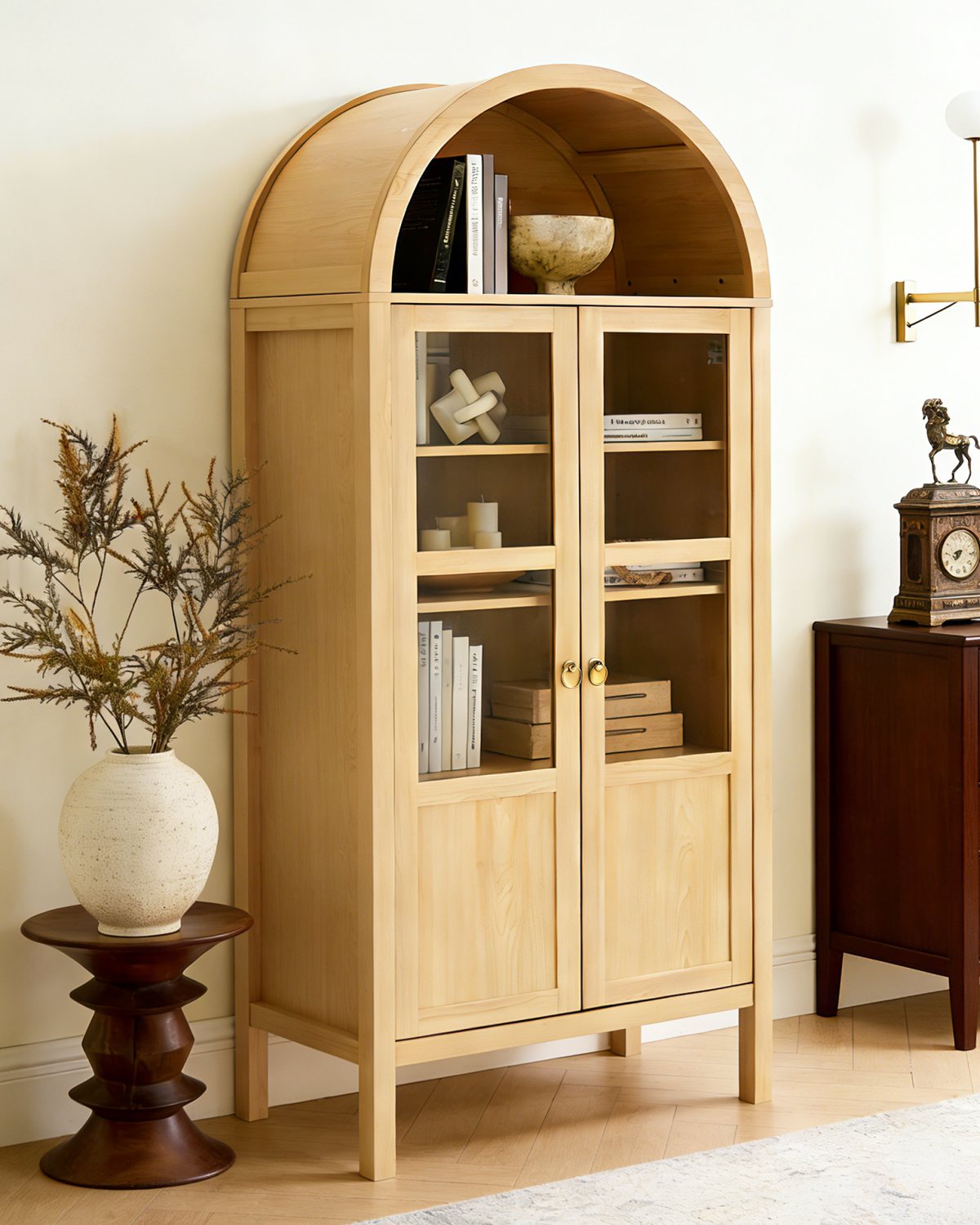 A tall arched cabinet with glass doors styled in a dining room, beside a plant stand and a dark wood chest.